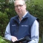 In einem Waldstück steht ein Mann mit Brille und blauer Weste über einem gestreiften Hemd im Freien. Er hält ein Notizbuch in der Hand und scheint in die Kamera zu blicken, vielleicht denkt er über Digitalisierungstrends für IT-EDEN nach. Bäume und Grünflächen sorgen für die ruhige Kulisse.