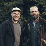 Zwei Männer stehen draußen vor Bäumen. Beide tragen Brillen und Mützen und legere Jacken. Der Mann links lächelt, während der Mann rechts einen Bart und einen neutralen Gesichtsausdruck hat.