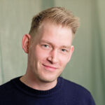 Eine Person mit kurzen hellen Haaren und dunklem Pullover lächelt leicht und blickt in die Kamera. Der Hintergrund ist in einem sanften, verschwommenen Grün gehalten.