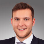 Ein Mann mit kurzen braunen Haaren und gestutztem Bart lächelt. Er trägt einen dunklen Anzug, ein weißes Hemd und eine Krawatte. Der Hintergrund ist ein neutraler Grauverlauf.