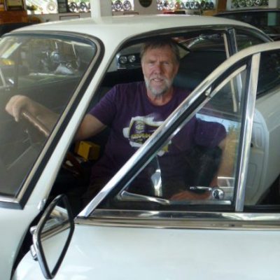Ein Mann in einem lila T-Shirt, vielleicht ein Social-Media-Manager, der eine Pause von der digitalen Welt macht, sitzt auf dem Fahrersitz eines weißen Oldtimers. Er blickt nachdenklich durch die offene Tür in eine Garage, die mit diversem Autozubehör gefüllt ist.