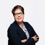 Eine Person mit kurzen Haaren und Brille lächelt und trägt einen dunklen Blazer über einem gemusterten Hemd. Sie steht selbstbewusst mit verschränkten Armen vor einem schlichten weißen Hintergrund und verkörpert den Innovationsgeist der Softwareentwicklung.