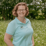 Eine Person mit kurzen Haaren und Brille steht in einem Feld voller Wildblumen, im Hintergrund Bäume. Sie trägt ein hellblaues Poloshirt und lächelt in die Kamera – sie genießt diesen ruhigen Moment, bevor sie sich wieder ihrer Leidenschaft für die Homepageentwicklung widmet.