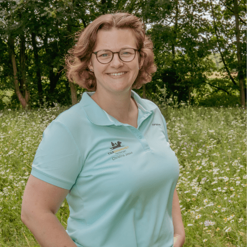 Eine Person mit kurzen Haaren und Brille steht in einem Feld voller Wildblumen, im Hintergrund Bäume. Sie trägt ein hellblaues Poloshirt und lächelt in die Kamera – sie genießt diesen ruhigen Moment, bevor sie sich wieder ihrer Leidenschaft für die Homepageentwicklung widmet.