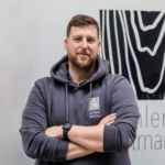 Ein bärtiger Mann, der einen dunklen Kapuzenpullover mit Logo und eine Apple Watch trägt, steht selbstbewusst mit verschränkten Armen vor einer mit abstrakten Designs geschmückten Wand und verkörpert die Essenz professionellen Online-Präsenz-Managements.