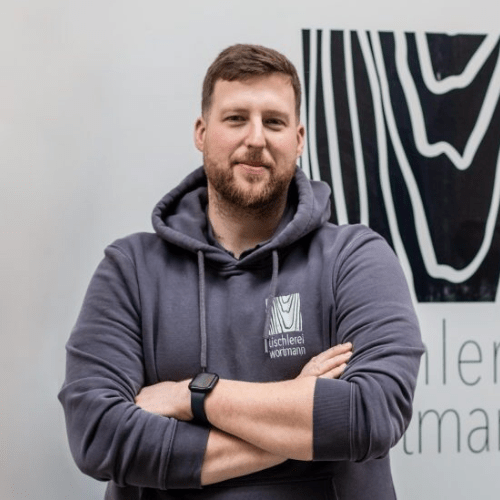 Ein bärtiger Mann, der einen dunklen Kapuzenpullover mit Logo und eine Apple Watch trägt, steht selbstbewusst mit verschränkten Armen vor einer mit abstrakten Designs geschmückten Wand und verkörpert die Essenz professionellen Online-Präsenz-Managements.